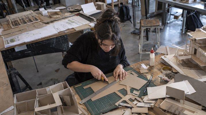 Una estudiante corta cartón para una maqueta en una mesa de taller llena de reglas y maquetas