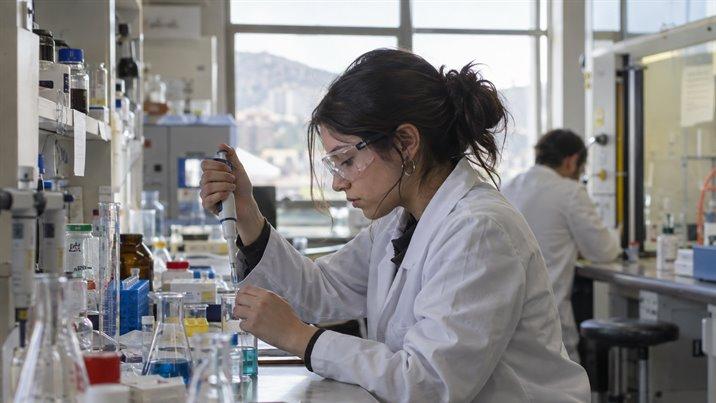 Una estudiante con delantal blanco y antiparras usa una pipeta en un laboratorio de química