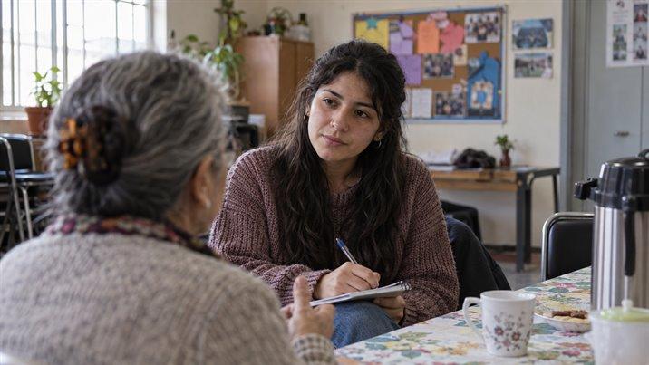 Una joven conversa con una señora mayor y toma notas en una libreta, en una sede vecinal