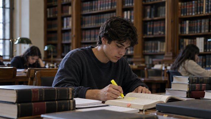 Un estudiante subraya un libro en una biblioteca antigua, con libros gruesos apilados a su lado