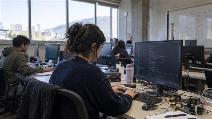 Una estudiante trabaja frente a un computador en un laboratorio, con una placa electrónica y cables en la mesa