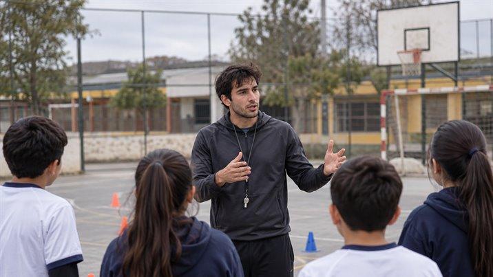 Un profesor con buzo deportivo y silbato explica un ejercicio a un grupo de escolares en una cancha
