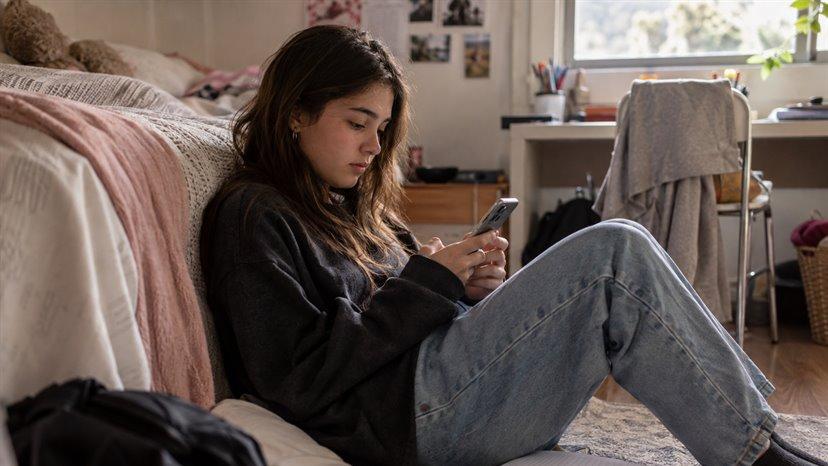 Una estudiante sentada en el suelo de su pieza, mirando el celular con un cuaderno al lado
