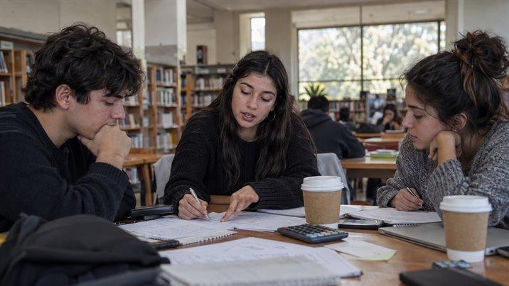Tres estudiantes trabajan en grupo en la mesa de una biblioteca, con apuntes y vasos de café