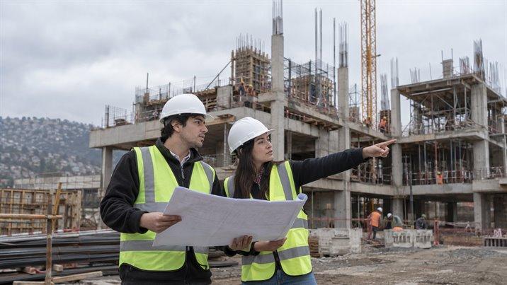 Dos jóvenes con casco blanco y chaleco reflectante revisan un plano en una obra en construcción