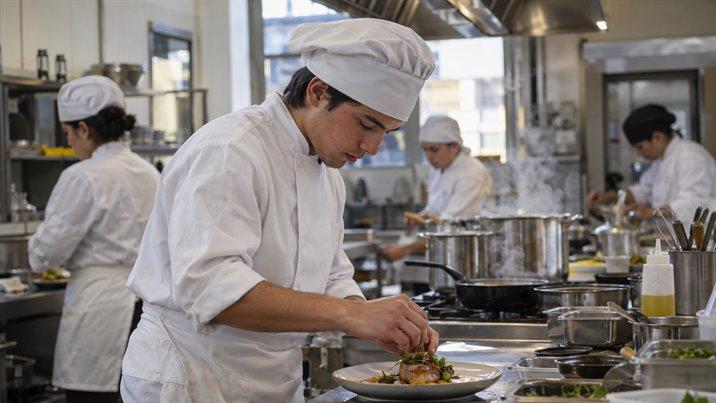 Un estudiante con chaqueta y gorro de cocinero emplata un plato en la cocina de una escuela