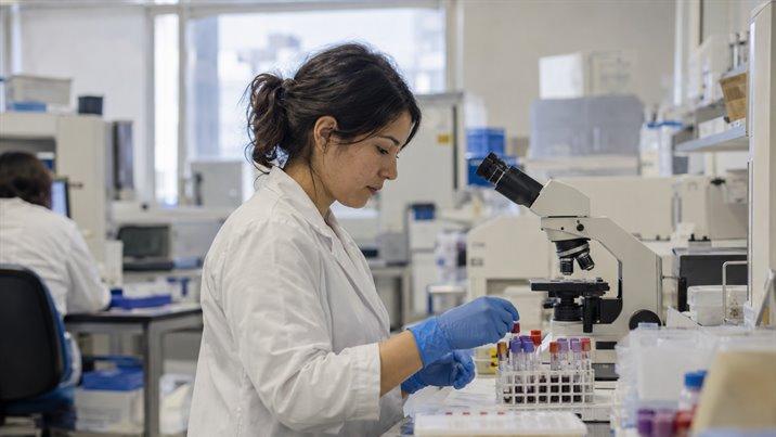 Una joven con delantal blanco y guantes ordena tubos de muestra junto a un microscopio en un laboratorio