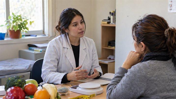 Una nutricionista conversa con una paciente en su consulta, con frutas y verduras sobre el escritorio
