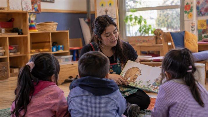 Una educadora sentada en el suelo de un jardín infantil les lee un cuento a tres niños pequeños
