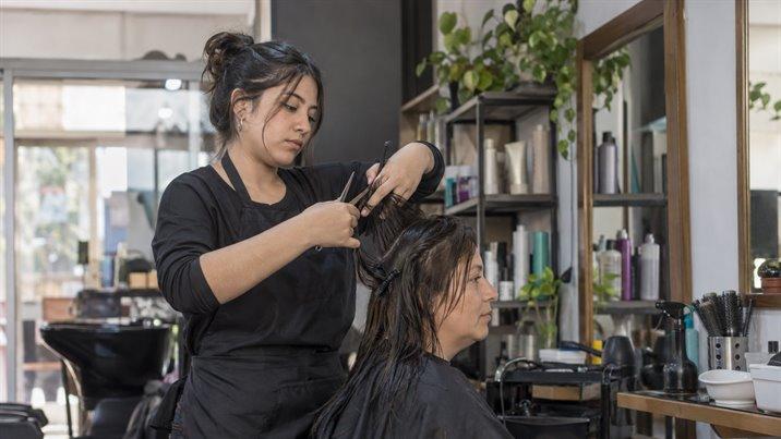 Una estudiante le corta el pelo con tijeras y peineta a una clienta sentada en una peluquería