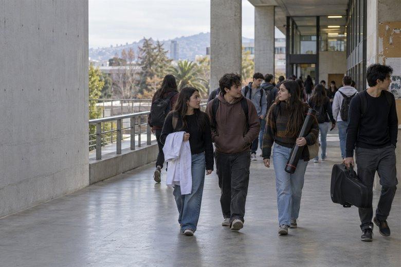 Estudiantes caminando por el pasillo de un instituto a la hora de salida, con mochilas y un tubo de planos